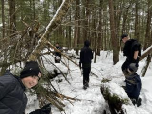 Students in winter gear walk through the snowy forest near partially built shelters and fallen branches.