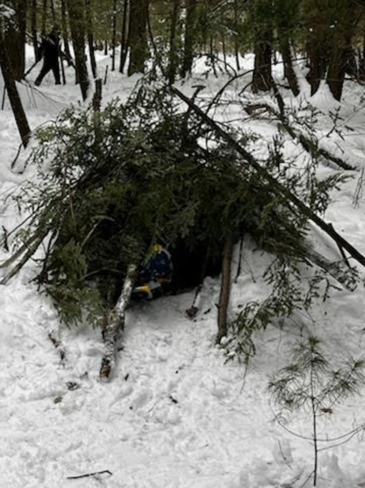 A simple lean-to style shelter made of branches and evergreen boughs built in the snow.