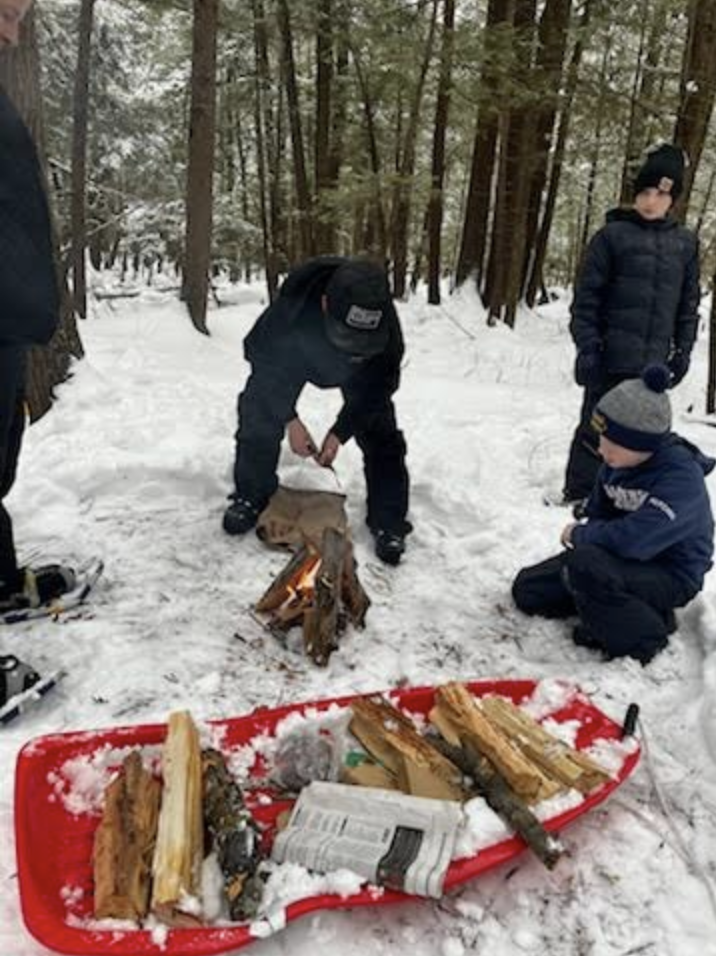 An adult helps students ignite a small campfire in the snow while others watch and kneel nearby.
