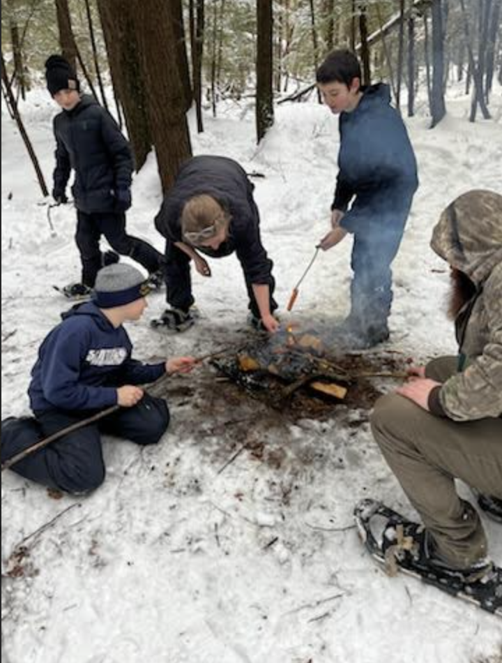 Small group of students around a campfire in the woods, roasting food on sticks over the flames.