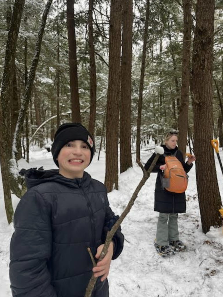Student in winter gear smiles while holding a stick; another person with a backpack stands behind among snow-covered trees.