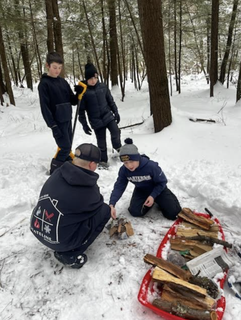 Students in winter clothing gather in the woods around a small fire setup, with a red sled full of firewood nearby.
