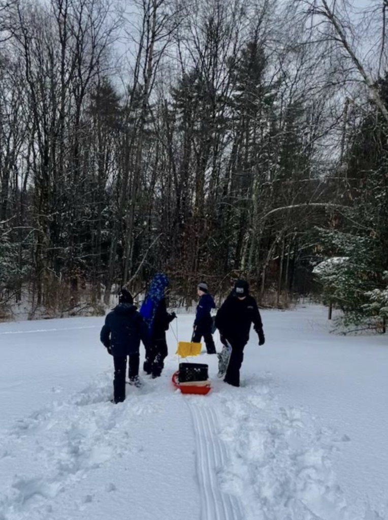 Group of students snowshoeing through a snowy field toward a wooded area, pulling a red sled with gear behind them.