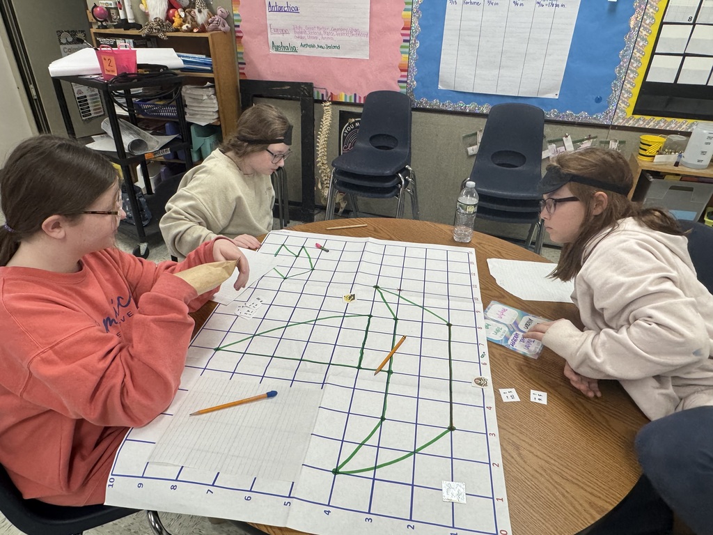 Two students collaborate on a coordinate grid activity at a round wooden table. One student, wearing an eyepatch on her forehead, holds an orange marker while the other student, wearing an eyepatch over her eye, leans over to connect points on the large grid paper. A brown envelope labeled "3" and several crayons sit nearby