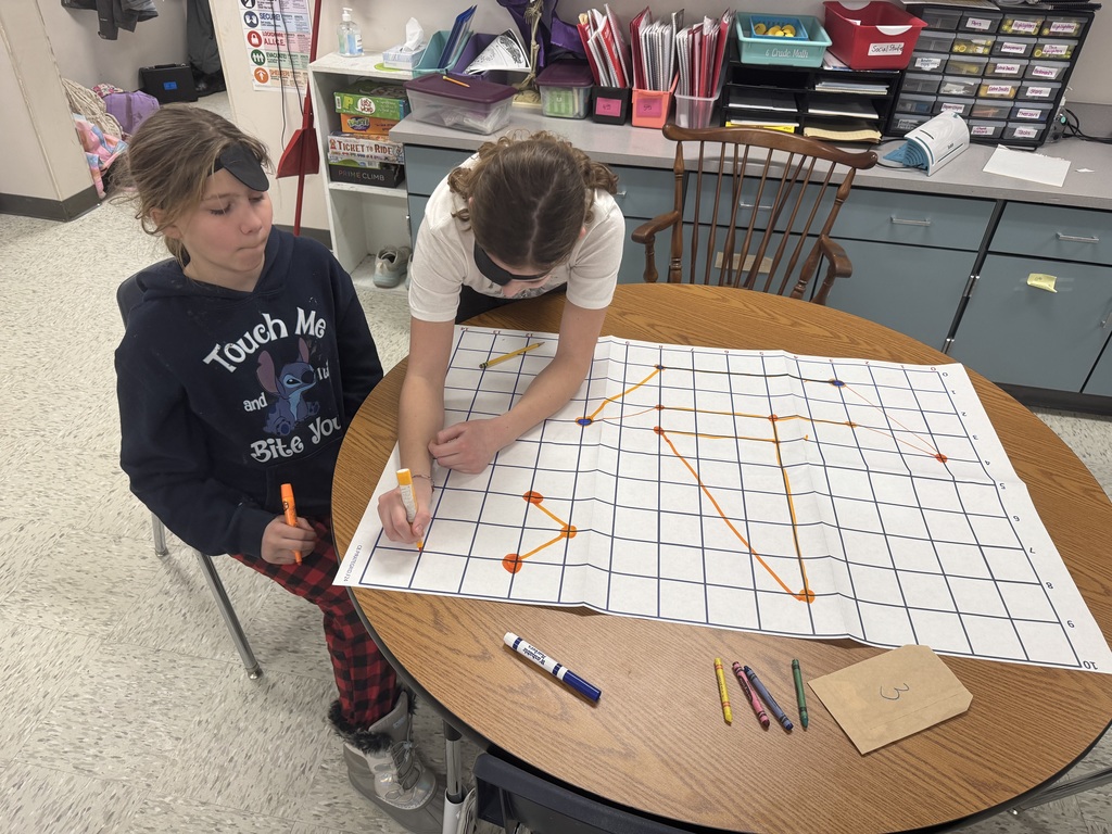 Gemini said Two students in a classroom work together on a large coordinate grid map spread across a round wooden table. One student, wearing an eye patch on her forehead, watches as the other draws orange lines connecting points on the grid. Various markers and a brown envelope labeled "3" are scattered on the table.
