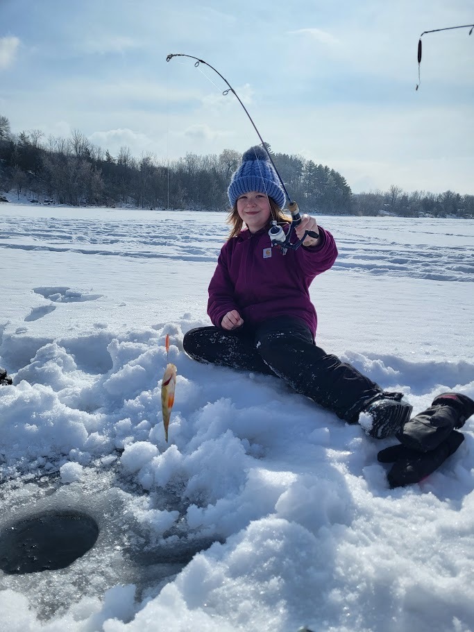A smiling student sitting on the ice holds up a fishing rod next to a freshly caught fish on the snow.