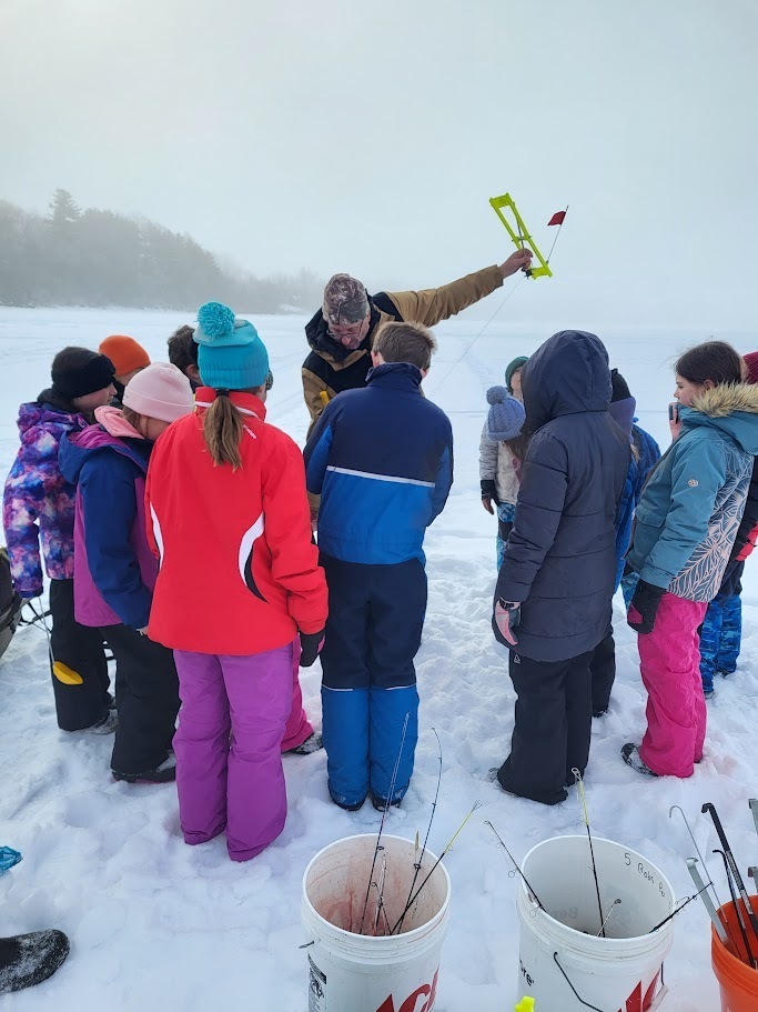 An instructor holds up an ice fishing tip-up while students in colorful winter clothing gather around to watch.