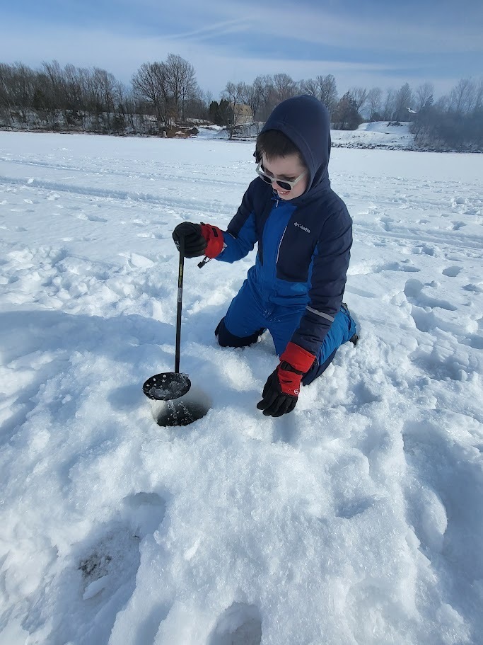 A student in winter gear kneels beside an ice hole using a skimmer to clear slush from the water.