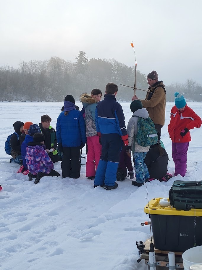 Students in winter gear gather on the ice as an instructor demonstrates ice fishing equipment next to sleds and supply bins.