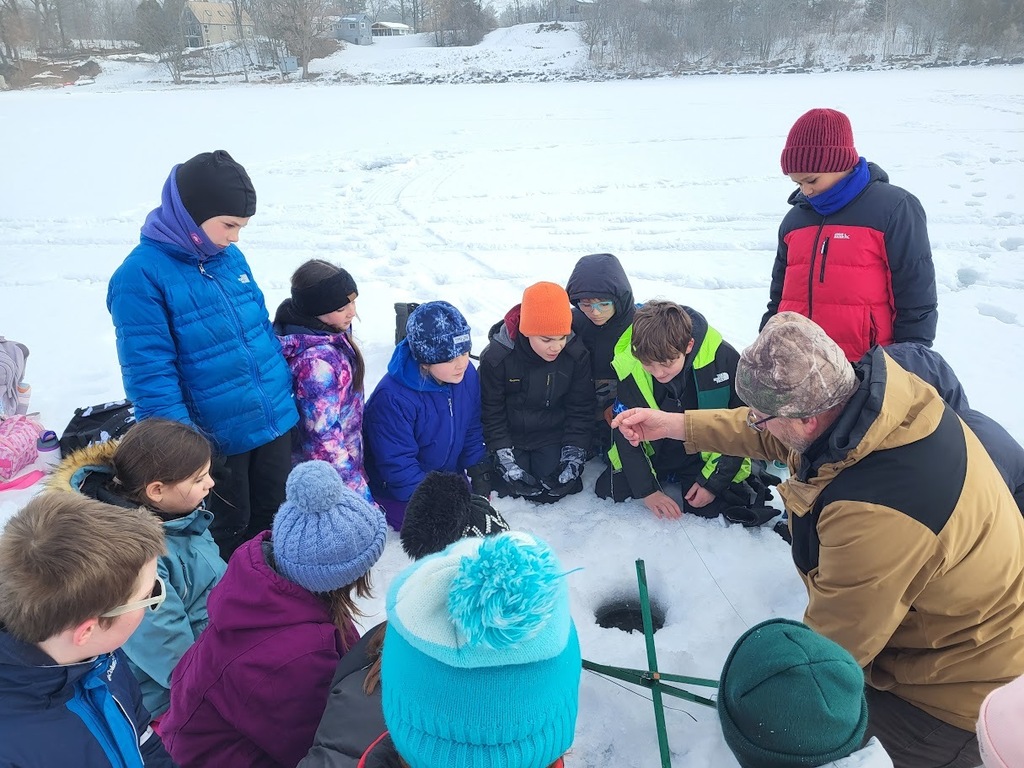 A group of bundled students kneels around an ice hole while an instructor shows them how to use ice fishing tools.