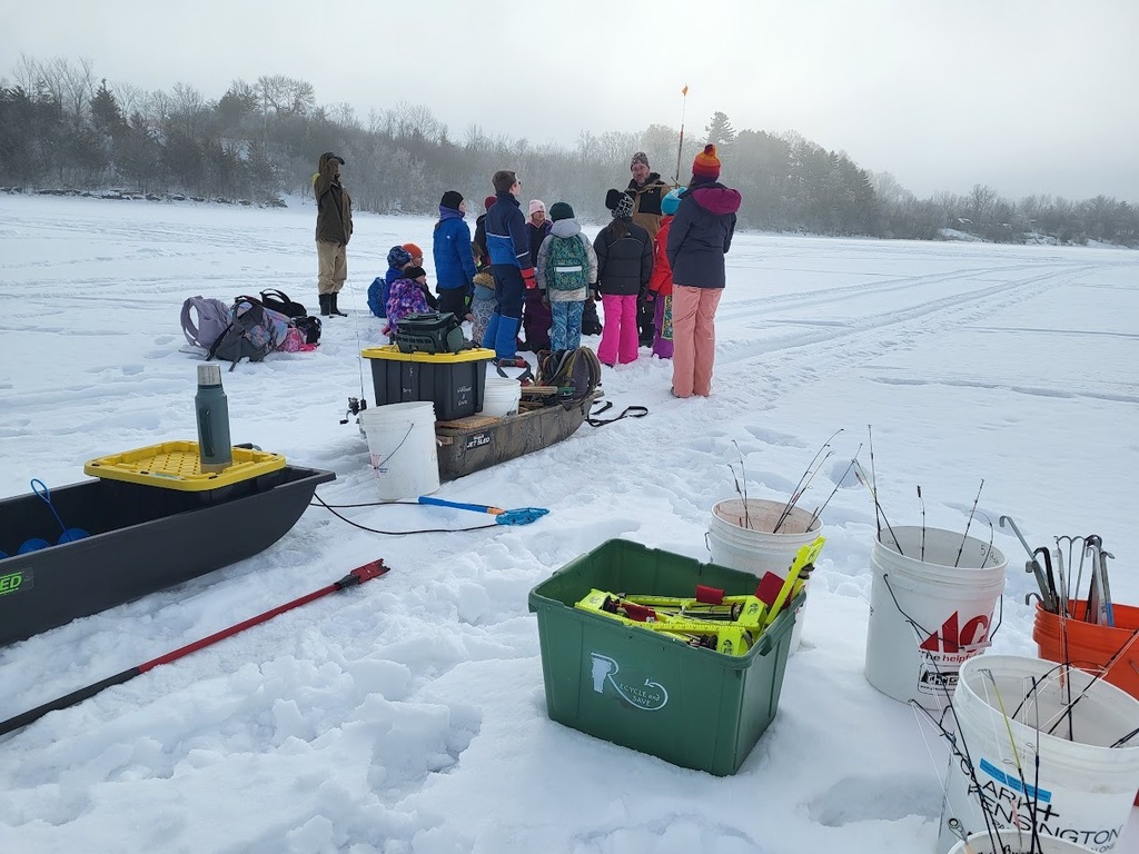 Ice fishing supplies—including buckets, rods, and sleds—sit on the frozen lake while students listen to instruction in the background.