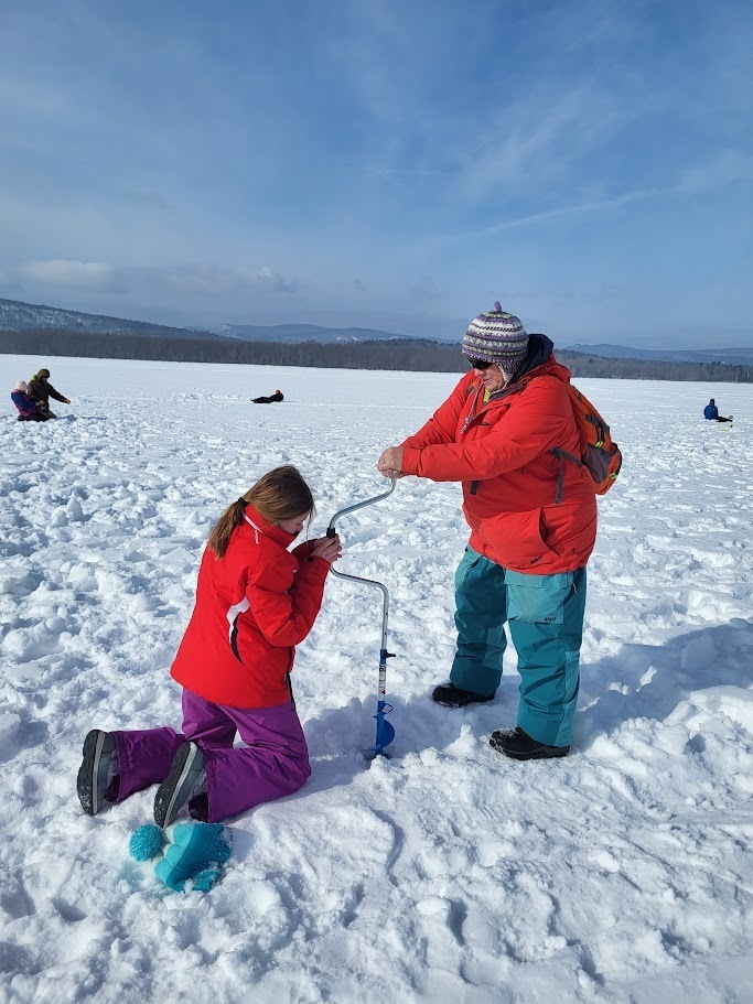 An adult helps a student operate an ice auger to drill a hole in the frozen lake during an outdoor learning activity.