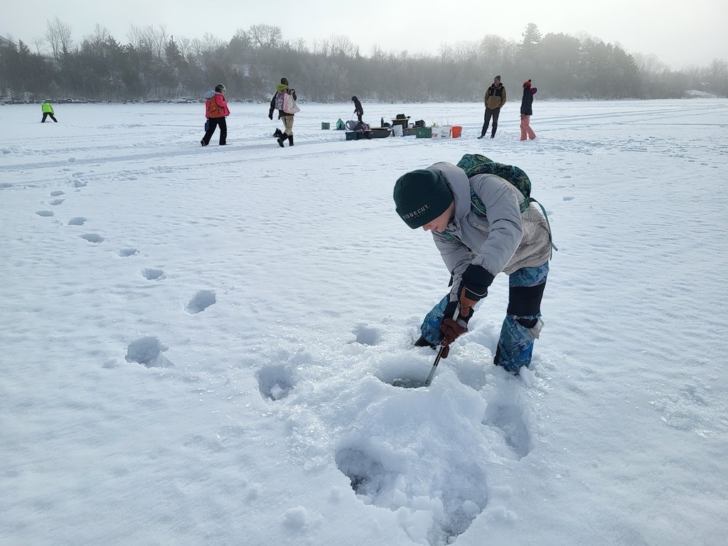 A student kneels on the ice and drills into a hole while classmates and adults stand nearby on the frozen lake.
