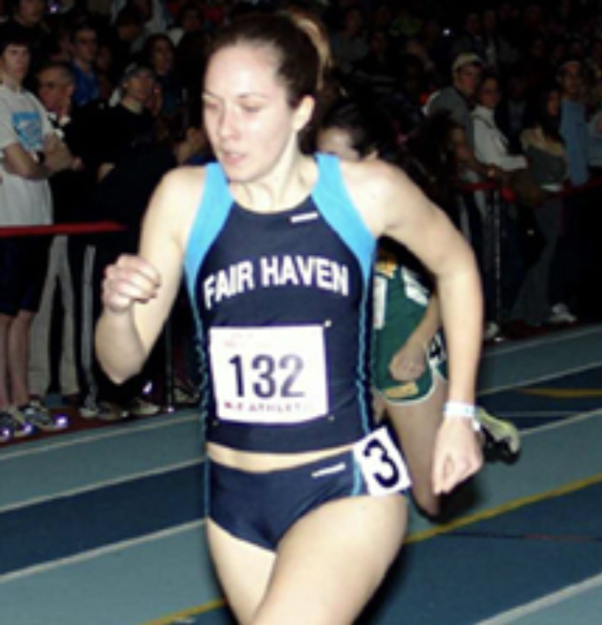 Student athlete in a Fair Haven track uniform runs on an indoor track during a race, wearing bib number 132, with spectators visible in the background.