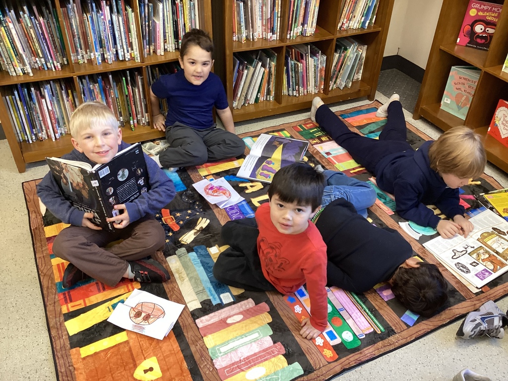 Five elementary students sit and lie on a colorful reading rug in the library, looking at books and drawings while surrounded by bookshelves.
