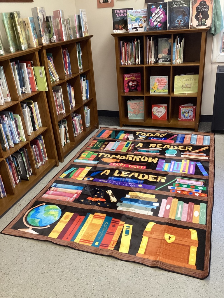 Colorful reading rug with illustrated bookshelves and the words “Today a Reader, Tomorrow a Leader” displayed in a school library corner surrounded by wooden bookshelves filled with children’s books.