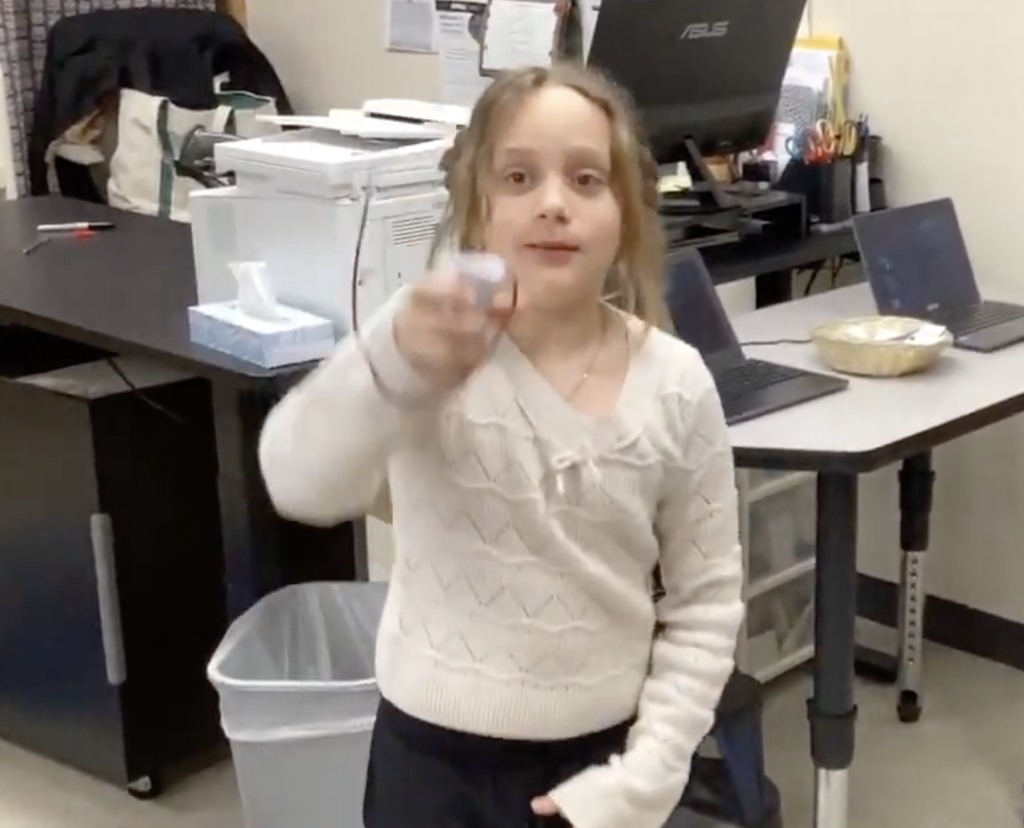 A student in a classroom holds up a small handmade basketball hoop game toward the camera, demonstrating the project after the class reading activity.