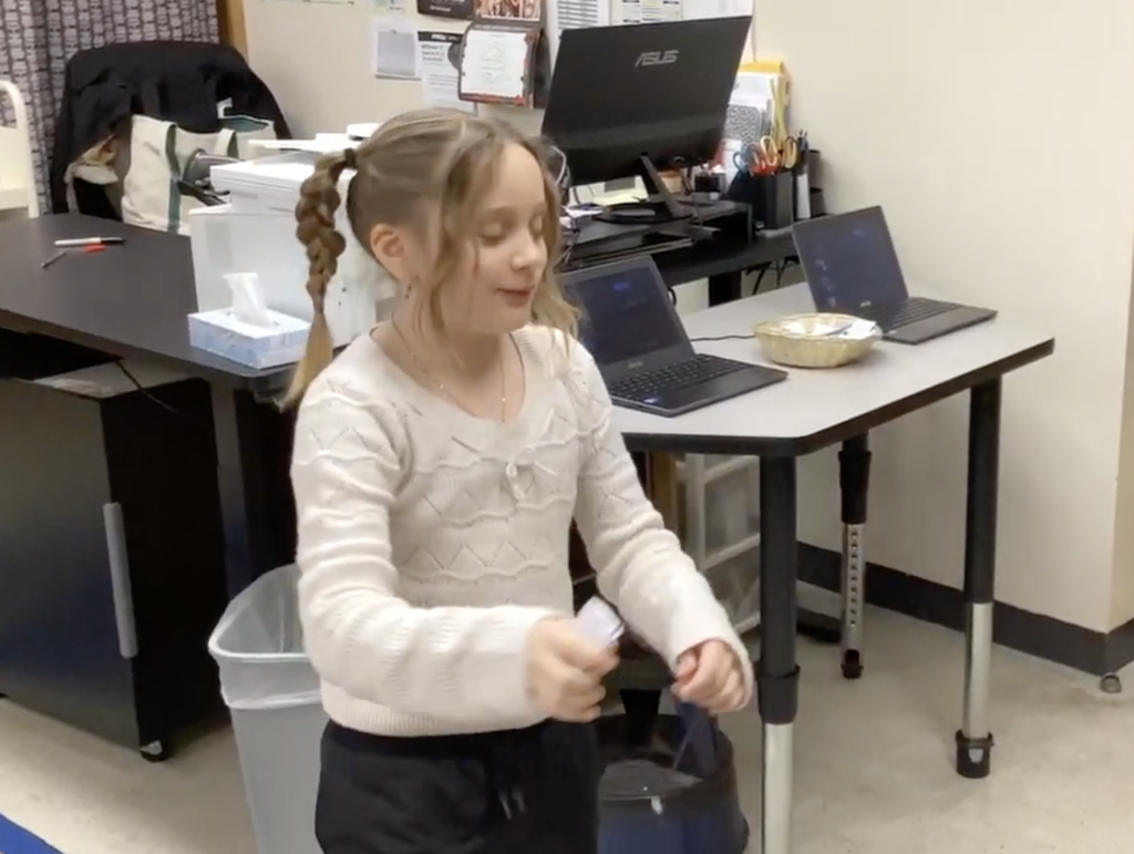 A young student in a classroom holds a small handmade basketball hoop game while standing near a table with laptops and a printer in the background.