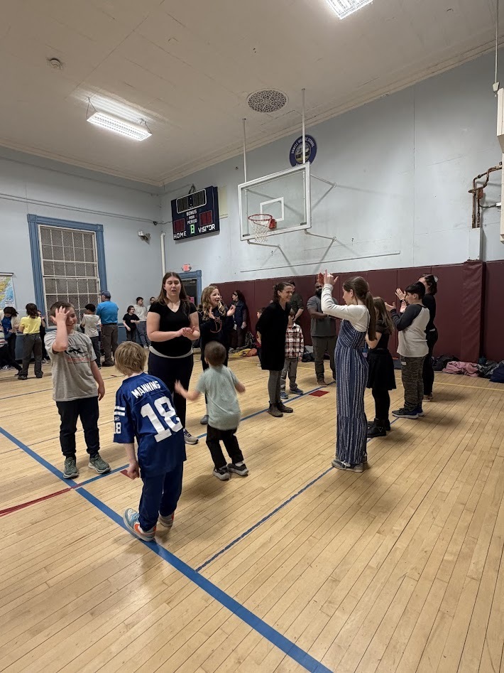 Students and adults form small dance groups in the gym, clapping and moving to the music during a lively school community event.