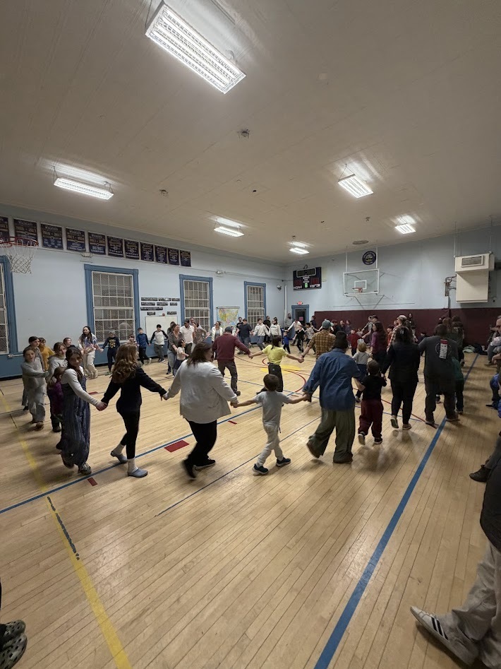Large group of students, staff, and families holding hands in a wide circle while dancing together in a school gymnasium.