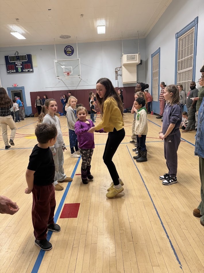 Older student leads a younger child in a dance line in the gym while other children and adults watch and wait their turn.