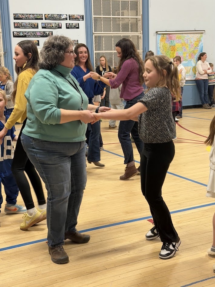 Adult and elementary student face each other and hold hands while dancing in a crowded school gym during a family event.