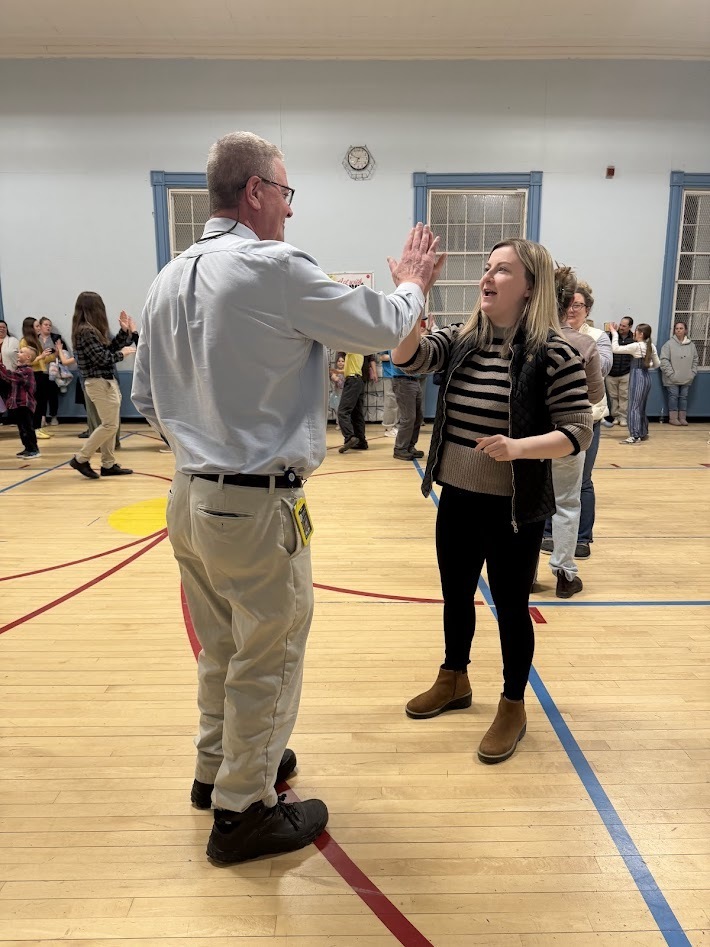 Two adults in a school gym smile and give each other a high five during a community dance event while others participate in the background.