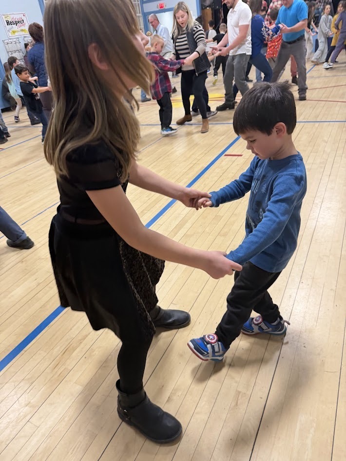 Young student and older student hold hands and practice dance steps together on the gym floor as other participants dance behind them.