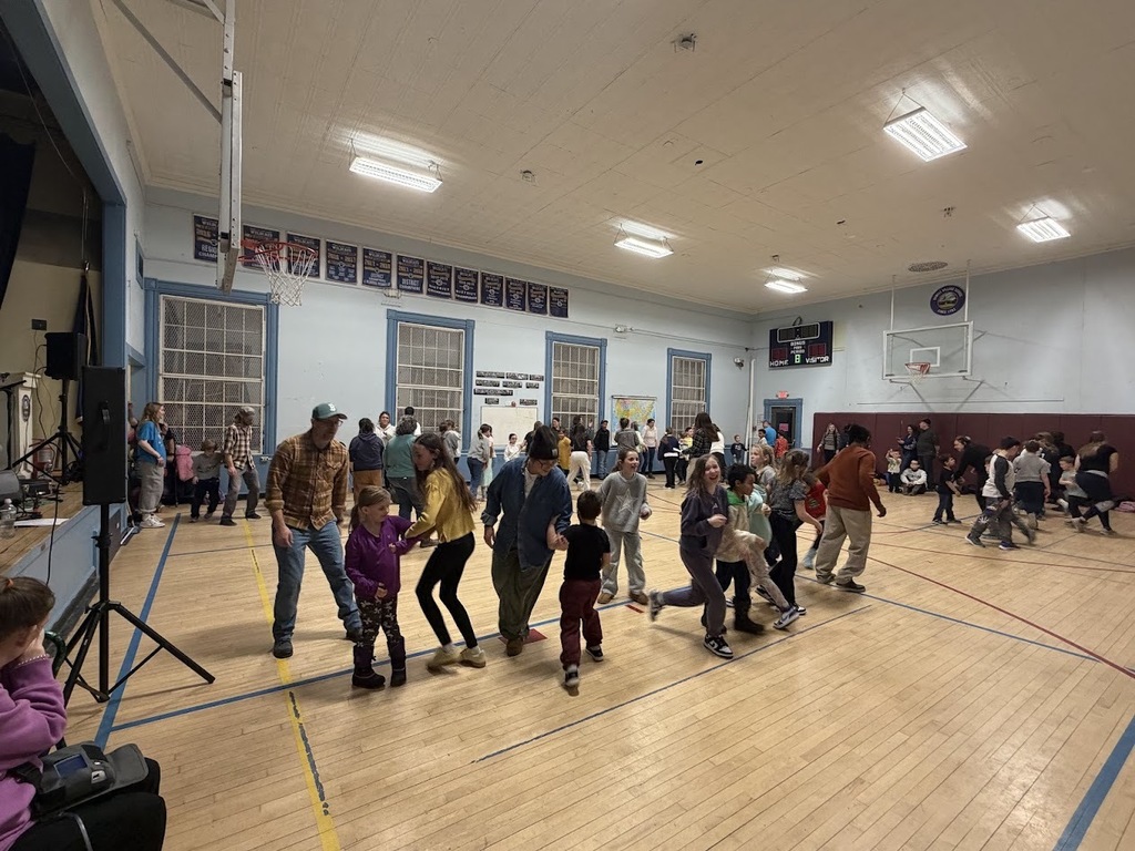Wide view of a busy school gym filled with families and students dancing together in lines and small groups.