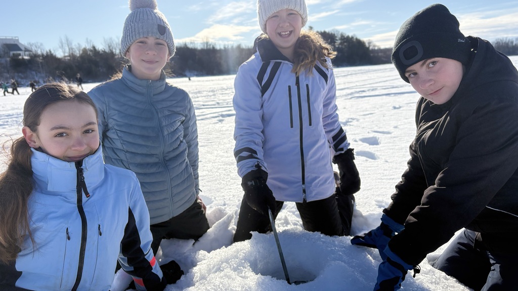 Four students in winter gear kneel together around an ice hole, smiling toward the camera on the frozen lake.