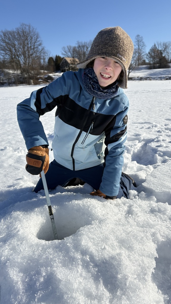 Student in a blue jacket and winter hat kneels on the ice using a metal tool to check an ice fishing hole.