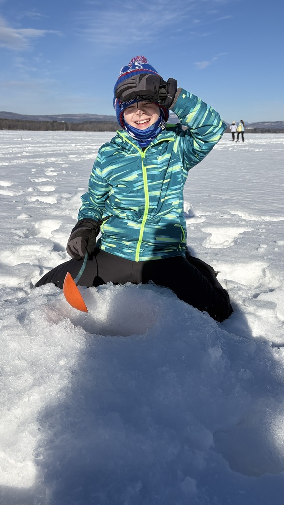 Smiling student in a blue and green winter jacket kneels beside an ice hole holding an ice fishing scoop on a sunny day.