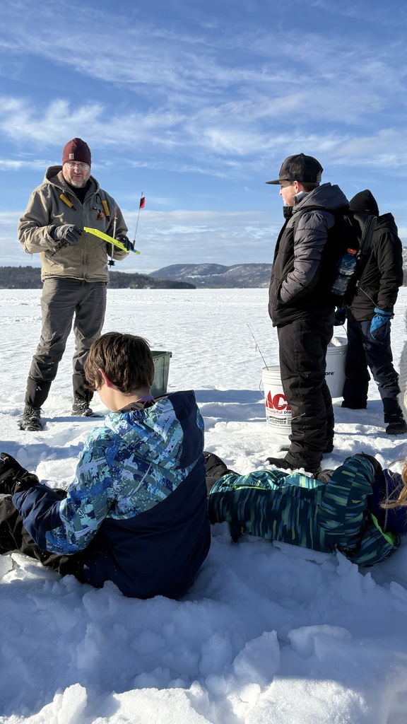 Adult instructor demonstrates ice fishing equipment to a group of bundled-up students gathered on the snowy ice with mountains in the background.