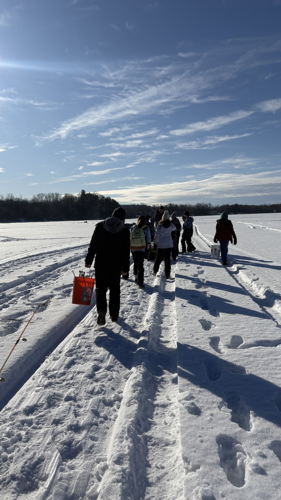 Group of students and adults walk in a line across a snowy frozen lake, carrying buckets and gear under a bright blue winter sky.