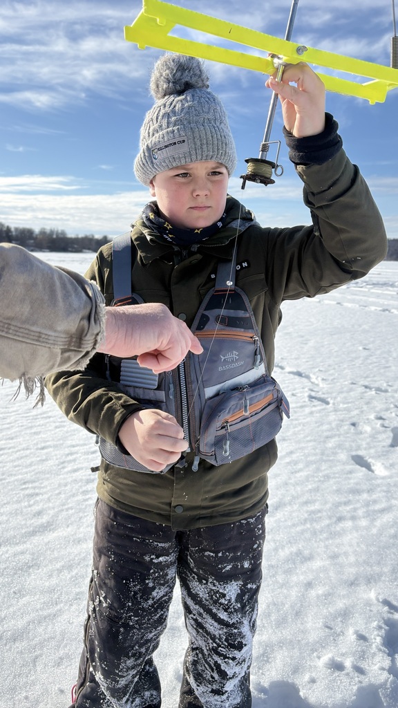 Student wearing a gray knit hat and winter bibs examines an ice fishing tip-up while an adult hand points to the line.
