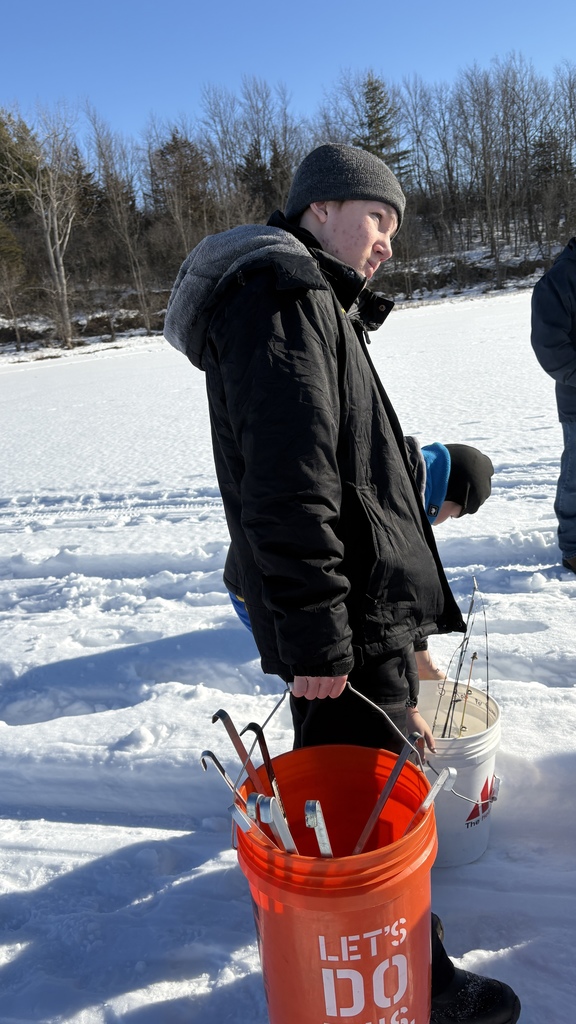 Student in a winter jacket and gray knit hat carries an orange bucket filled with ice fishing tools while standing on the frozen lake.