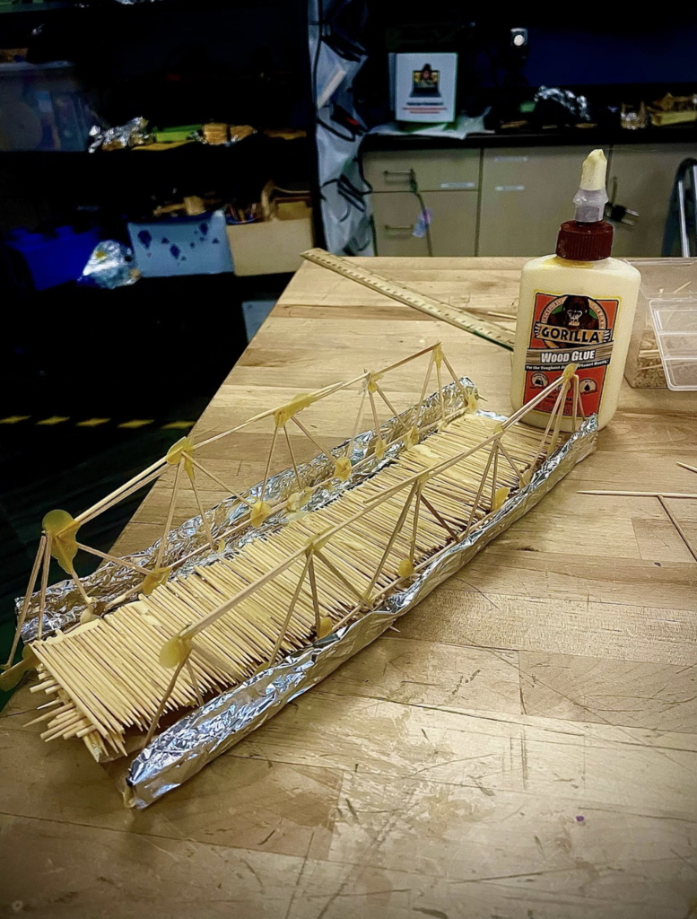 Completed toothpick suspension-style bridge sits on a workbench next to a bottle of wood glue.