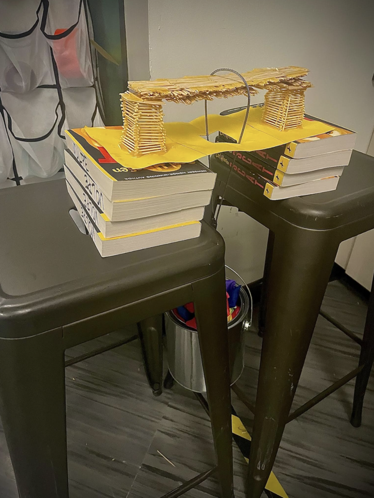 Close-up of a toothpick bridge resting on stacks of books between two stools during a weight test setup.