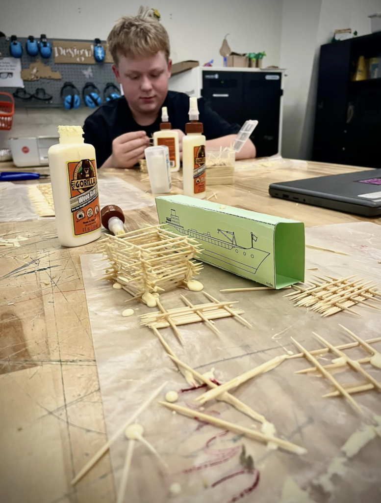Student concentrates while gluing toothpicks together on a classroom desk.
