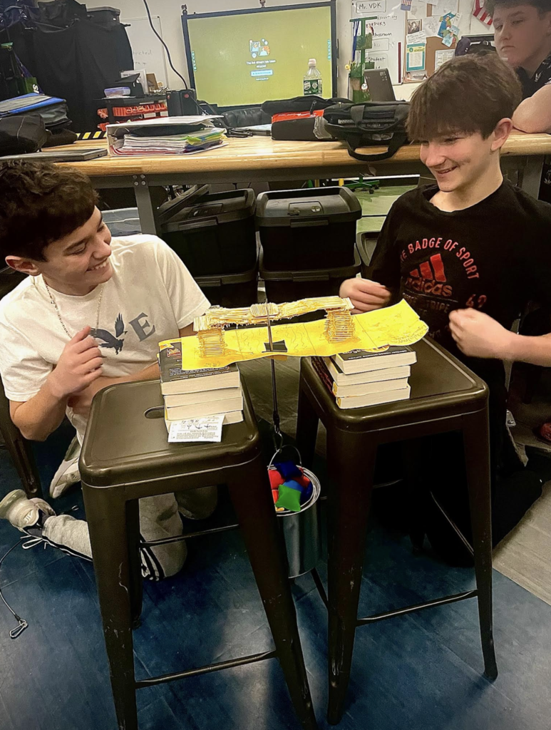 Two middle school students smile while testing a yellow paper and toothpick bridge supported between two stools with weights hanging below.