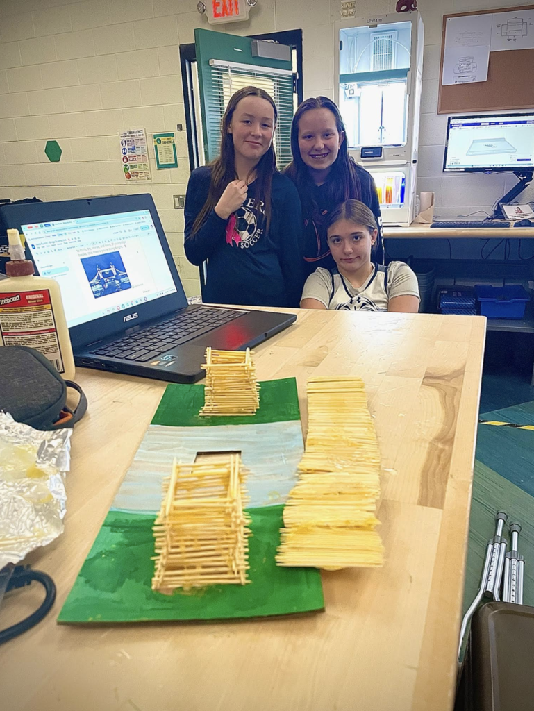 Three students pose behind their finished toothpick bridge structures on a classroom table.
