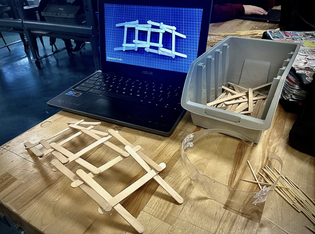 Laptop displays a bridge design reference while craft sticks and materials are spread across a classroom worktable.