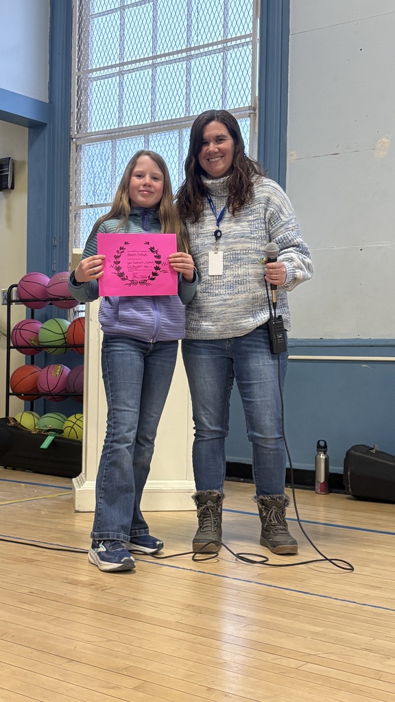 Student stands next to a staff member in the gym, holding a bright pink certificate during a school recognition moment.