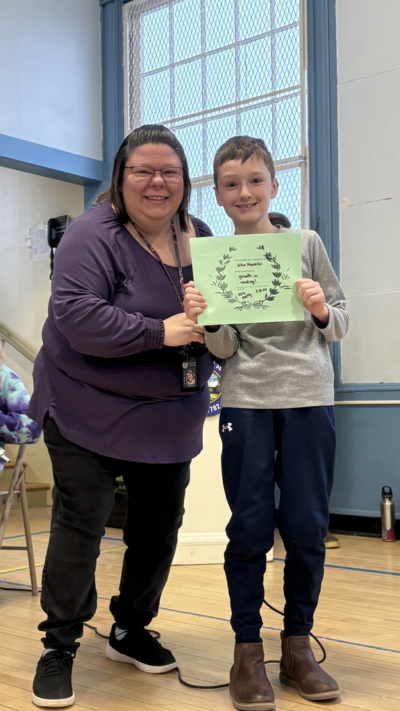 Smiling student stands with a staff member in the gym, holding a green certificate recognizing growth in reading.