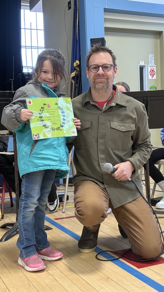 Young student stands beside an adult holding a microphone in the school gym, proudly displaying a green award certificate.
