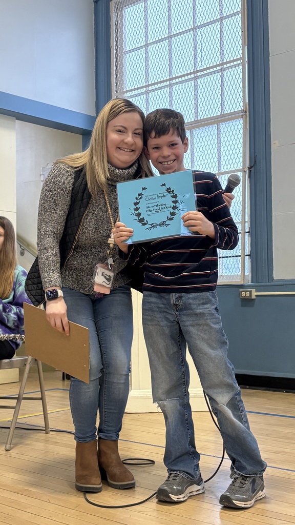 Student and teacher pose together in the gym, holding a blue award certificate while the student also holds a microphone.