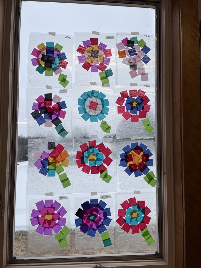 A large window with twelve colorful tissue paper flower artworks taped to the glass in a 3-by-4 grid. Each flower is made from overlapping translucent squares in bright colors like pink, purple, blue, green, and red, with green stems. Snowy landscape and bare trees are visible outside through the window.