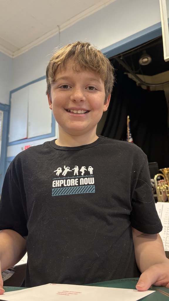 Student in a black “Explore Now” T-shirt smiles while holding a green folder and Student of the Month envelope.