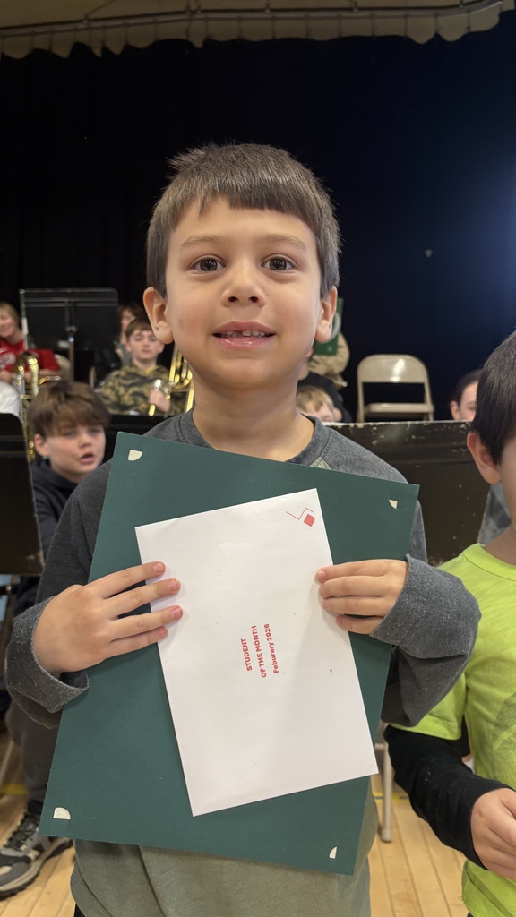Elementary student holds a green certificate folder with a white “Student of the Month February 2026” envelope, standing in front of seated band students.