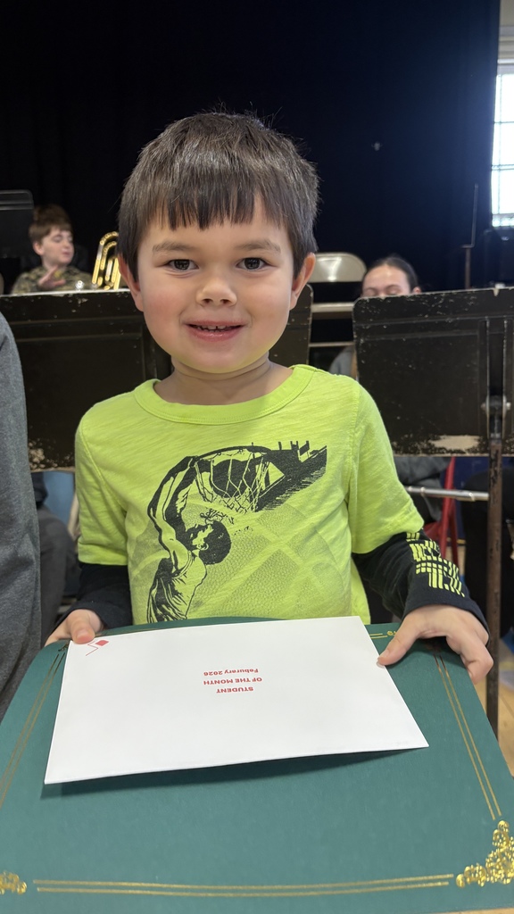Young student in a bright green shirt smiles while holding a green folder and a white “Student of the Month February 2026” envelope in front of the school band area.